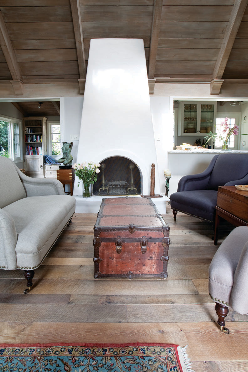 A cozy living room featuring reclaimed wood flooring with rich grain patterns and a weathered finish. The antique-style space is complemented by vintage decor, a white plaster fireplace, and natural light, highlighting the warmth and character of the reclaimed flooring